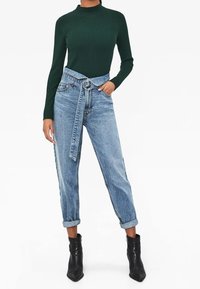 Grünes geripptes Langarmoberteil, kombiniert mit hellblauen high-waisted Jeans mit gebundenem Gürtel und aufgerollten Bündchen, ergänzt durch schwarze Knöchelstiefel.