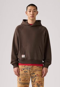 Giovane uomo che indossa una felpa marrone con tasca frontale e pantaloni beige con stampe e testo in stile cowboy occidentale, in piedi su uno sfondo semplice.