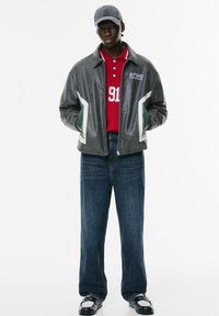 Giacca bomber in pelle grigia con accenti bianchi, polo rossa con il numero 91, jeans blu e scarpe nere. Gli accessori includono un cappellino grigio.