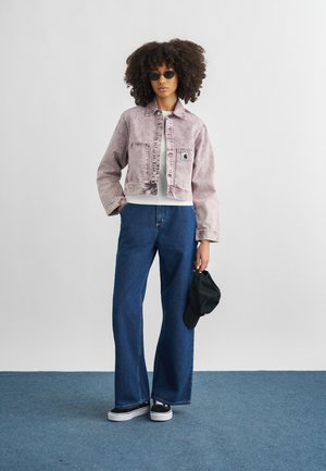 Femeie cu păr creț purtând ochelari de soare, jachetă roz pal, cămașă albă, blugi largi albaștri, adidași negri, ținând o șapcă neagră în mâna dreaptă.