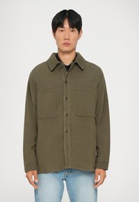Giacca camicia in corda verde oliva con chiusura a bottoni, due tasche sul petto e colletto a punta, indossata sopra una maglietta nera e jeans blu chiaro.