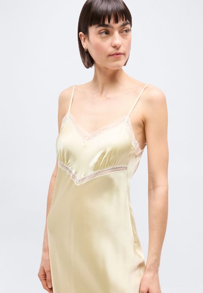 Γυναίκα που φοράει ροζέ χρυσό σατέν φόρεμα με λεπτομέρειες δαντέλ ας, κοιτάζοντας ελαφρώς προς τα δεξιά της, πάνω σε απλό φωτεινό φόντο.