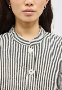 Γυναίκα που φοράει μια μαύρη και λευκή κάθετα ριγέ μπλούζα με μανσέτα μανταρινιού και δύο λευκά κουμπιά, με ασημένια σκουλαρίκια κρίκους.