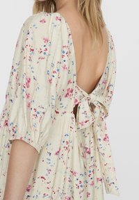 Žena oblečená do krémových květinových šatů s nafouklými rukávy a odhaleným zadním dílem, který je svázán mašlí.