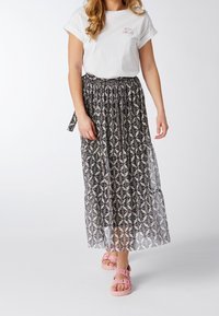 Mujer vestida con una camiseta blanca con texto, una falda midi de patrón en blanco y negro, y sandalias rosas caminando frente a un fondo liso.