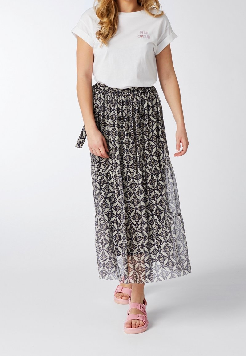 Mujer vestida con una camiseta blanca con texto, una falda midi de patrón en blanco y negro, y sandalias rosas caminando frente a un fondo liso.