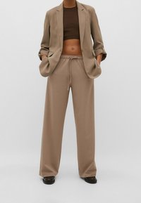 Blazer beige avec revers de manches retroussés, porté avec un crop top marron et un pantalon beige ample avec une taille élastique. Tissu à texture lisse.
