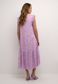 Lila cvetna obleka z nabranimi rokavi, slojevim krilom in majhnimi rumenimi ter roza cvetnimi vzorci. Obuta v sivkaste sandale s peto.