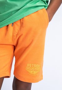 Oranje katoenen shorts met zijzakken en een gele logo tekst "PETROL INDUSTRIES" op de linkerdij. Groene shirt zichtbaar bovenaan.