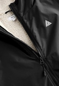 Black zippered jacket with a soft cream fleece interior, featuring a triangular logo and a smooth outer texture. Zipper detail is prominent.