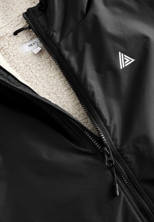 Black zippered jacket with a soft cream fleece interior, featuring a triangular logo and a smooth outer texture. Zipper detail is prominent.
