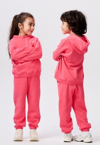 Twee kinderen dragen bijpassende roze hoodies en broekjes, met elastische manchetten en een losse pasvorm, met witte sneakers.
