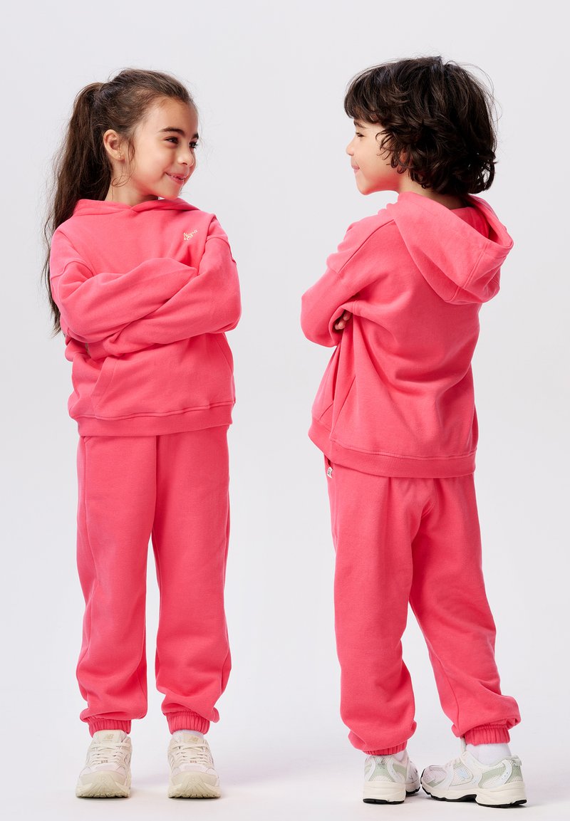 Twee kinderen dragen bijpassende roze hoodies en broekjes, met elastische manchetten en een losse pasvorm, met witte sneakers.
