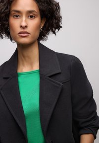 Femme aux cheveux bouclés foncés portant des boucles d'oreilles en perle, un haut vert et un manteau noir aux manches retroussées sur un fond uni.