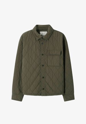 Jonge man staat met handen in de zakken, draagt een olijfgroene gewatteerde jas, een wit-groen gestreept shirt, bruine ribfluwelen broek en donkerbruine schoenen.