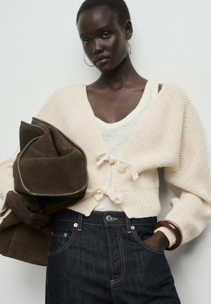 Cárdigan de punto acanalado color crema con cierres de lazo, superpuesto sobre un top blanco. También son visibles unos jeans de mezclilla oscura y un bolso de ante marrón.