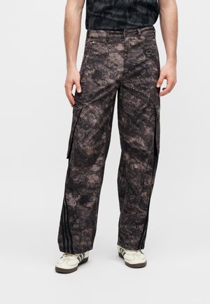 Homme portant un pantalon cargo ample à motifs foncés avec des poches latérales et des baskets blanches sur un fond uni.