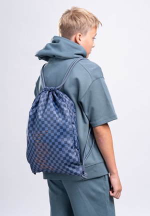 Ragazzo giovane in felpa con cappuccio verde acqua e pantaloni, con uno zaino a coulisse a quadri blu con testo arancione, di profilo su uno sfondo semplice.