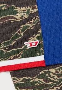Pletena tkanina z teksturiranim kamuflažnim vzorcem v zelenih in rjavih odtenkih, s trdnimi modrimi, rdečimi in belimi akcenti; vsebuje majhen vezen logotip.