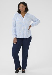 Blouse à manches longues bleu clair avec un motif paisley blanc, décolleté en V et détails de boutons, associée à un jean droit bleu foncé et des chaussures noires.