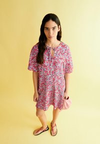 Blumendruckkleid in rosa und lila Tönen mit lockerer Passform, Schlüsselloch-Ausschnitt und kurzen Ärmeln. Getragen mit schwarzen Flip-Flops und einer pinken Tasche.