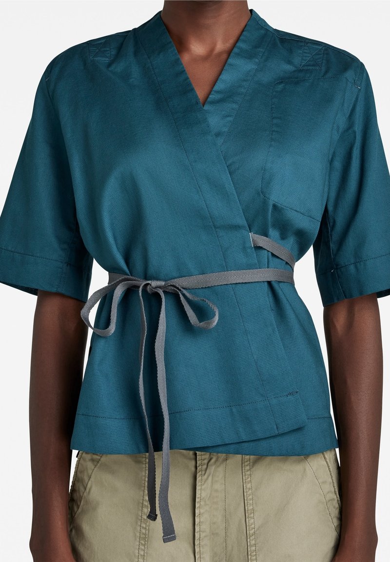 Haut court à manches courtes de style drapé en sarcelle avec ceinture grise à la taille, porté avec un pantalon vert olive clair sur une personne à la peau foncée.