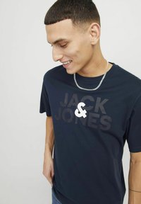 Tumesinise puuvillane T-särk, millel on silmapaistev musta ja valge "JACK & JONES" logo. Lühikesed varrukad ja lõdvestunud lõige. Näha on hõbedane kett. 
