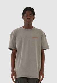 Overdimensioneret grå bomulds-t-shirt med korte ærmer, med et lille brunt grafisk motiv på brystet og en klassisk rund halsudskæring.