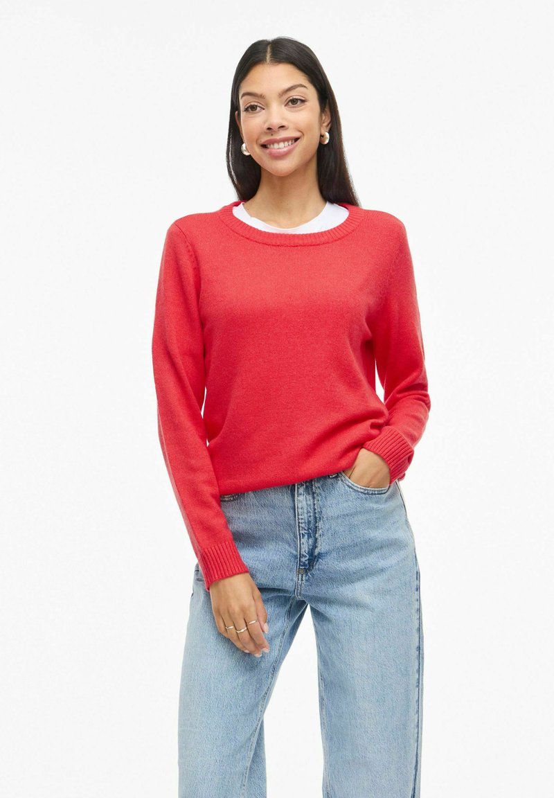 Femme aux longs cheveux bruns portant un pull rouge, un jean bleu clair et des boucles d'oreilles créoles, souriant avec une main dans la poche.