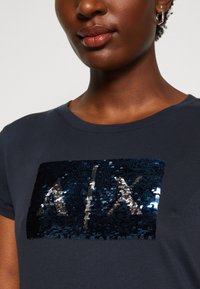 Mørkeblå T-skjorte med et firkantet sequinmotiv med et blankt, mønstrete design. Rund hals og korte ermer. Myk tekstur.