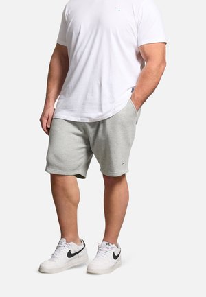 Homme portant un t-shirt blanc, un short gris clair et des baskets blanches avec le logo Nike noir, debout avec une main dans sa poche.
