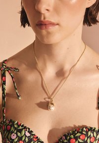 Zelta ķēdes kaklarota ar apaļu pērles piekariņu un nelieliem dekoratīviem akcentiem, nēsāta virs ziedu raksta, volānu rotātas blūzes.