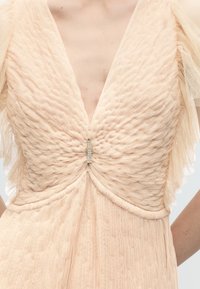 Κοντινό πλάνο σε μπεζ υφασμάτινο φόρεμα με βαθύ V-λαιμόκοψη, ραμμένο μπούστο, διακριτικά διάφανα φουσκωτά μανίκια και μικρό κούμπωμα με στρας στο κέντρο.