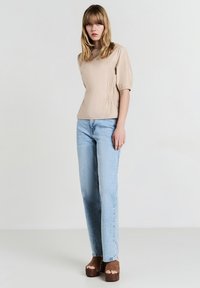 Blusa beige con maniche a sbuffo, dettaglio arricciato sui lati, abbinata a jeans dritti blu chiaro. Tacchi a piattaforma marroni completano il look.