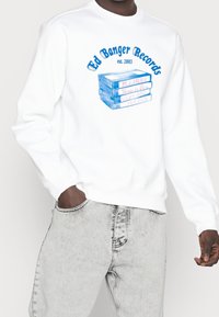 Persona che indossa una felpa bianca con la scritta blu "Ed Banger Records est. 2003" e cassette impilate etichettate con titoli musicali.
