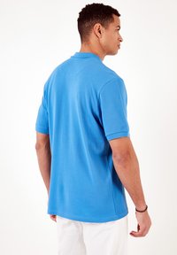 Camisa polo azul de manga corta hecha de un tejido texturizado. Presenta un cuello con solapa y un corte recto. La parte trasera es lisa y sin patrones visibles.