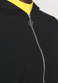 Felpa nera con zip realizzata in tessuto morbido, con una zip silver, una tasca frontale e un cappuccio. Sotto, è visibile un colletto giallo.