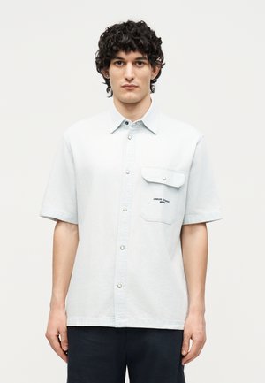 Joven con cabello rizado y oscuro lleva una camisa de botones azul claro de manga corta con bolsillo en el pecho, de pie frente a un fondo blanco liso.