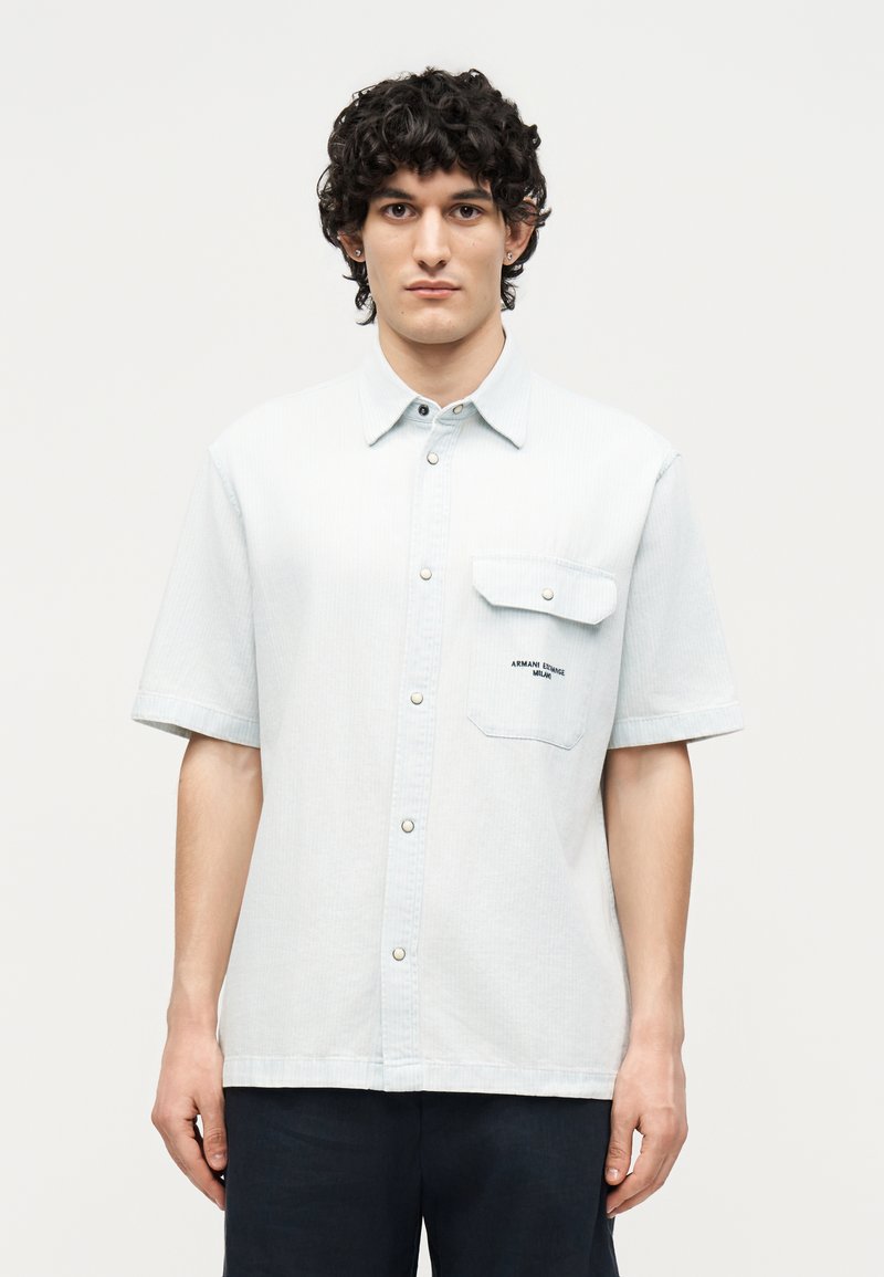 Νέος άνδρας με σγουρά σκούρα μαλλιά φοράει ανοιχτό μπλε κοντομάνικο πουκάμισο με τσέπη στο στήθος, στέκεται μπροστά σε απλό λευκό φόντο.