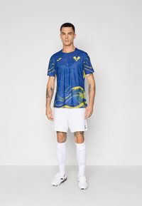 Maglia sportiva blu con accenti gialli e motivi a onde, abbinata a pantaloni corti bianchi e calzini sportivi bianchi. Modello che indossa scarpini da calcio.
