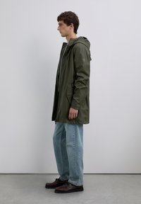 Ung man som står på sidan iförd en olivgrön regnjacka med huva, ljusblå jeans och mörkbruna lädercasualskor.