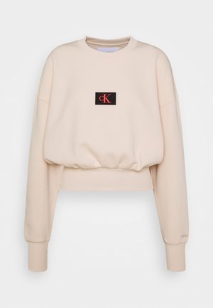 Sweatshirt beige raccourci en tissu doux, orné d'un patch rectangulaire noir avec "cK" en rouge. Manches côtelées et col rond.