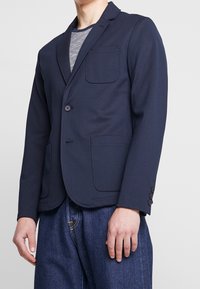 Námořnický blejzr z pružné látky, s klopovým límcem, jedním knoflíkem, náprsní kapsou a bočními kapsami. Pod ním je texturované šedé tričko.