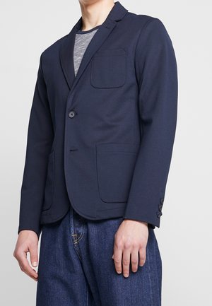 Marinblazer i stretchy tyg, med slagkrage, enkel knappstängning, bröstficka och sidofickor. Strukturerad grå T-shirt under.