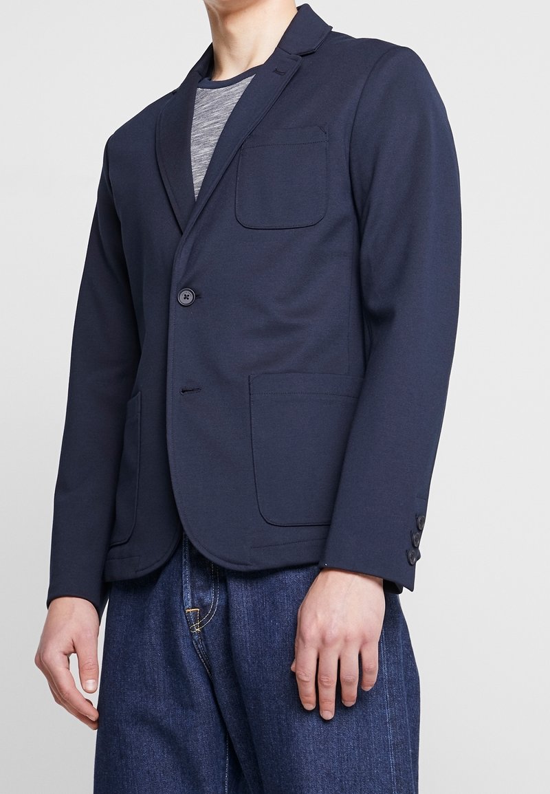 Námořnický blejzr z pružné látky, s klopovým límcem, jedním knoflíkem, náprsní kapsou a bočními kapsami. Pod ním je texturované šedé tričko.