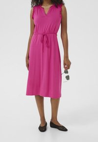 Robe rose sans manches avec des accents de volants aux épaules, décolleté en V, taille cintrée et longueur midi, accompagnée de ballerines en maille noires et de lunettes de soleil.