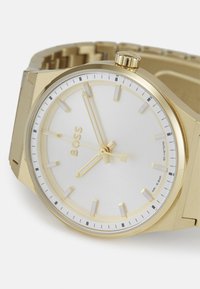 Reloj de acero inoxidable dorado con una esfera blanca, que incluye marcadores de hora y agujas doradas. Cuenta con una pulsera texturizada y una corona prominente.