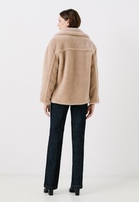Chaqueta de piel sintética en color beige claro, con un cuello ancho y un corte oversized, combinada con pantalones de mezclilla oscuros y zapatos de tacón negros.