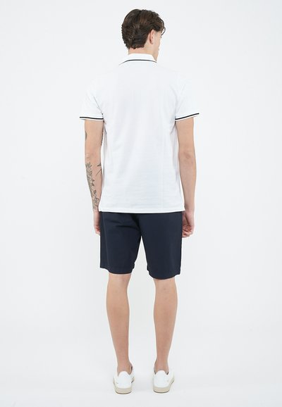 Mężczyzna stojący tyłem, ubrany w biały polo z czarnym wykończeniem, ciemne szorty, białe trampki oraz widoczne tatuaże na ramionach.