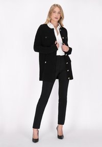 Schwarze Strickjacke mit zwei vorderen Taschen, goldfarbene Knöpfe, getragen über einem weißen Hemd und schwarzen Hosen, kombiniert mit schwarzen High Heels.